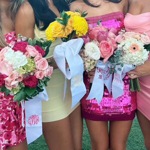 May include: Several bouquets of flowers with white ribbons. The bouquets feature a variety of flowers in shades of pink, yellow, and white. The ribbons have gold text. The women are wearing dresses in shades of pink and yellow.
