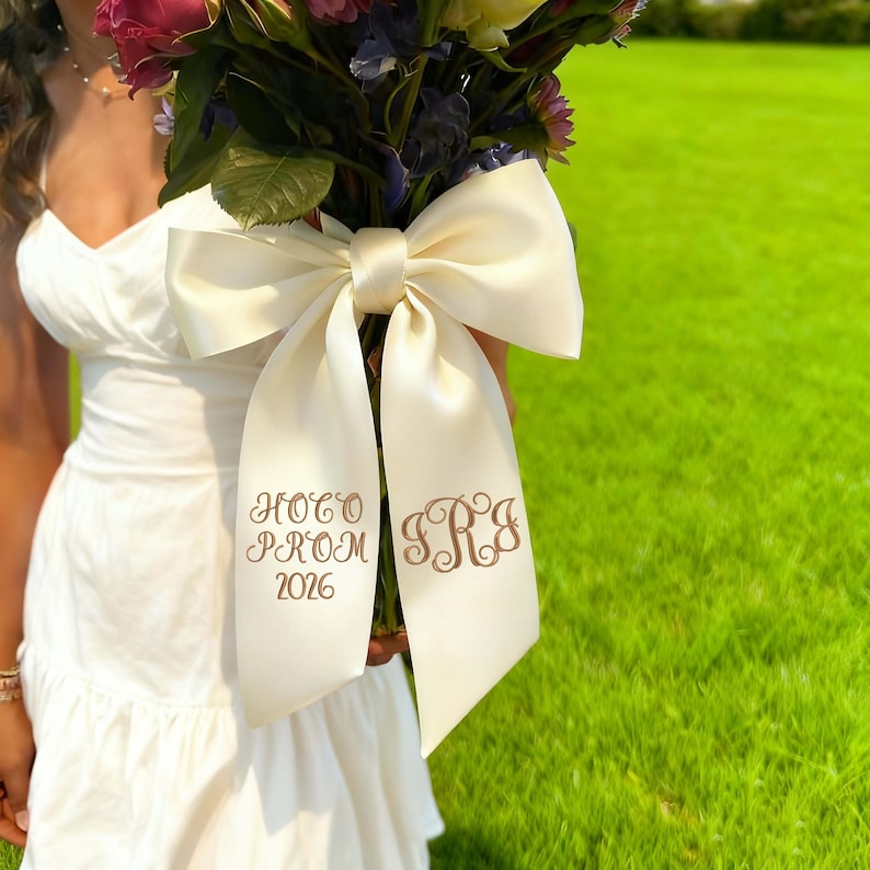 May include: A bouquet of flowers with a large ivory satin bow. The bow has the text "Hoco Prom 2026" and initials embroidered in a brown thread. The flowers are a mix of colors, including pink, purple, and white.