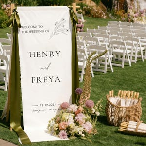 May include: A white wedding welcome sign with the text "WELCOME TO THE WEDDING OF HENRY and FREYA 12.12.2025". The sign is decorated with floral accents and olive green ribbons. A basket of parasols and floral arrangements are in front.