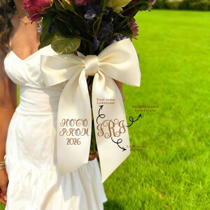 May include: A bouquet of flowers with a large ivory satin bow. The bow has custom embroidery with the words "Prom 2026" and initials. The background is a green lawn.