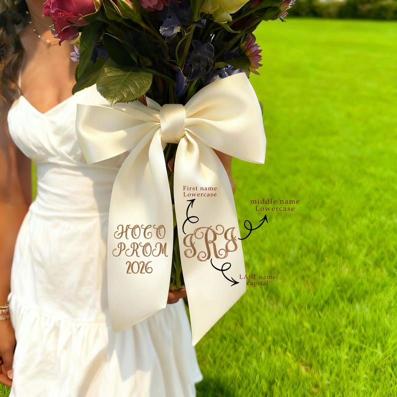 May include: A bouquet of flowers with a large ivory satin bow. The bow has custom embroidery with the words "Prom 2026" and initials. The background is a green lawn.