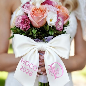Könnte beinhalten: Ein Hochzeitsstrauß mit rosa, pfirsichfarbenen und weißen Rosen und grünem Laub. Die Stiele sind mit einer weißen Satinschleife umwickelt, bestickt mit "HOCO 2026" und einem rosa Blumenmonogramm.