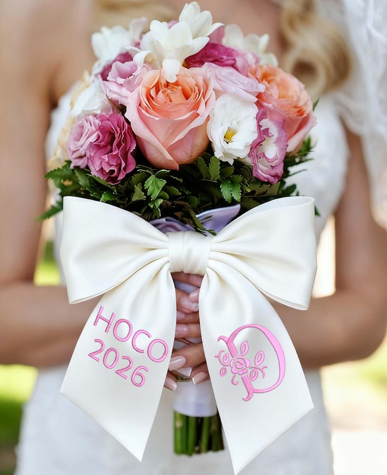 May include: A bridal bouquet featuring a mix of pink, peach, and white roses, with greenery. A large white satin bow is wrapped around the stems, embroidered with "HOCO 2026" and a pink floral monogram.