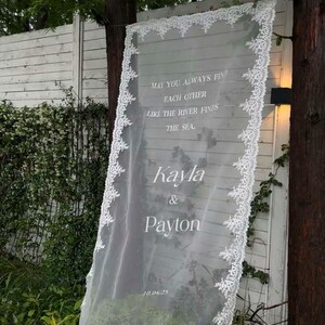 Pode incluir: Um painel de tecido branco transparente com delicadas bordas de renda, exibindo os nomes "Kayla & Payton" e a data "10.04.25." O painel apresenta a cita&ccedil;&atilde;o "May you always find each other like the river finds the sea."