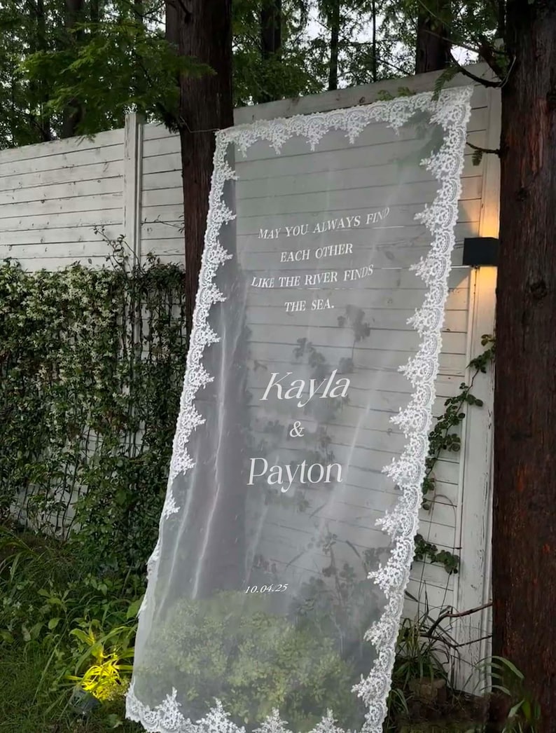 Pode incluir: Um painel de tecido branco transparente com delicadas bordas de renda, exibindo os nomes "Kayla & Payton" e a data "10.04.25." O painel apresenta a cita&ccedil;&atilde;o "May you always find each other like the river finds the sea."