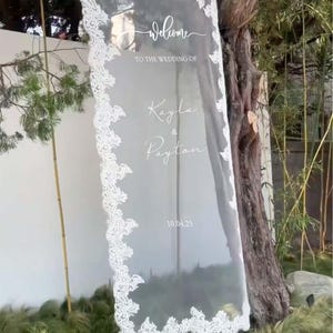 Pode incluir: Uma placa de boas-vindas de casamento em acr&iacute;lico transparente com acabamento em renda branca. A placa diz "welcome to the wedding of Kayla & Payton" e a data "10.04.25". A placa &eacute; exibida ao ar livre perto de vegeta&ccedil;&atilde;o e uma &aacute;rvore.