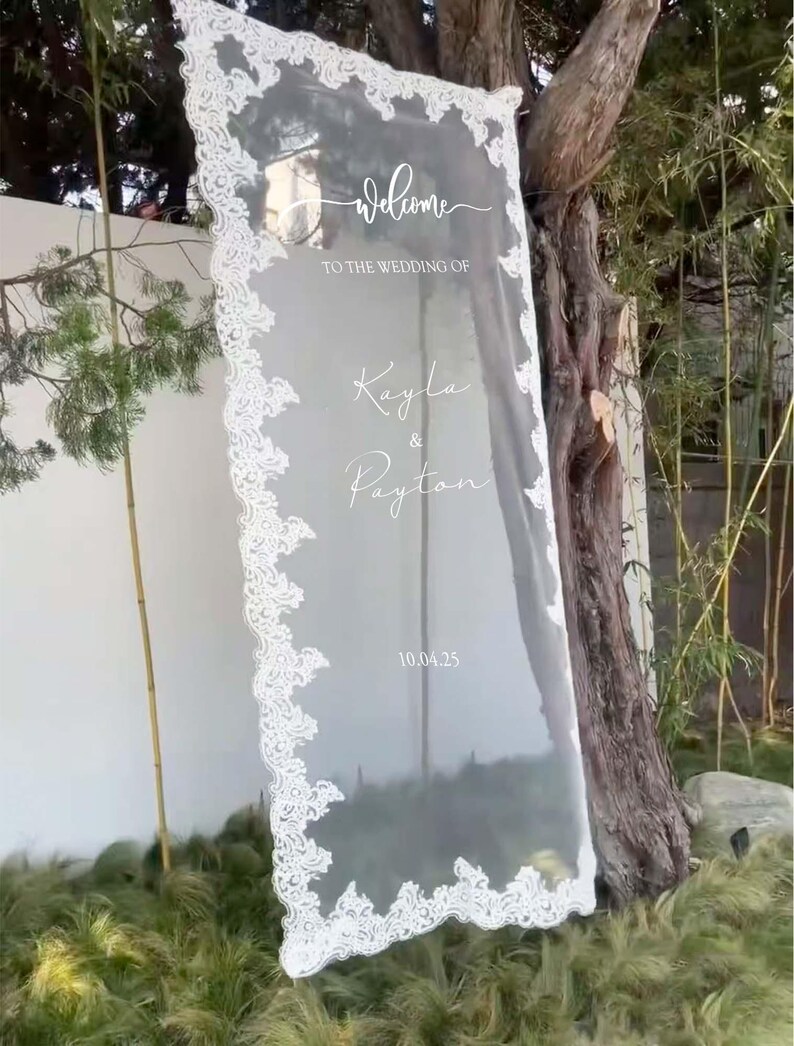 Pode incluir: Uma placa de boas-vindas de casamento em acr&iacute;lico transparente com acabamento em renda branca. A placa diz "welcome to the wedding of Kayla & Payton" e a data "10.04.25". A placa &eacute; exibida ao ar livre perto de vegeta&ccedil;&atilde;o e uma &aacute;rvore.