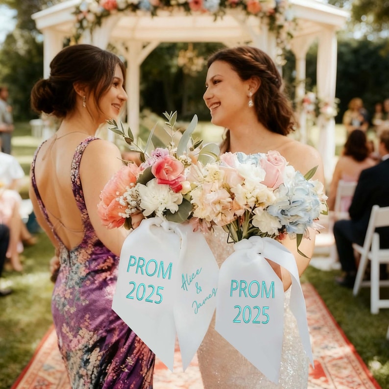 May include: Two women smiling, holding bouquets of flowers with white ribbons. The ribbons read "PROM 2025" in teal lettering, with one ribbon also displaying "Alice & James". The bouquets feature pink, white, and blue flowers. The women are in formal dresses.