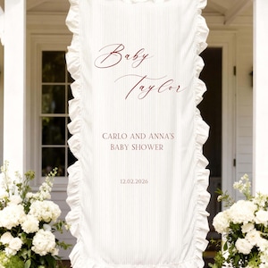 Può includere: Striscione in tessuto bianco con bordi a balze, che mostra il testo "Baby Taylor" in elegante scrittura. Sotto, si legge "Carlo and Anna's Baby Shower" e la data 12.02.2026. Lo striscione è appeso davanti a una casa bianca con composizioni floreali.