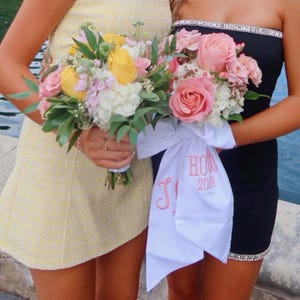 May include: Two bouquets of flowers, one with yellow and pink roses, the other with pink roses. The bouquet with pink roses has a white ribbon with the text "HON 2024" in pink. The person on the left is wearing a yellow dress, and the person on the right is wearing a black dress.