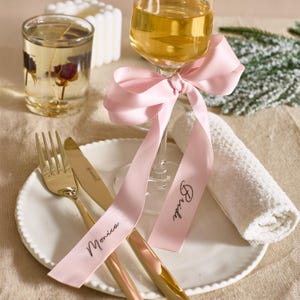 May include: A table setting with a white plate, gold cutlery, and a glass of wine adorned with a pink ribbon. The ribbon has the name "Monica" and "Bride" written on it. A small rolled white towel and a candle are also present.