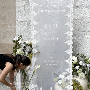 Pode incluir: Uma placa de boas-vindas de casamento com borda de renda branca e o texto "WELCOME TO THE WEDDING OF AXEL and KEILY". A data "18.10.2025" e o local "Peekskill, NY" tamb&eacute;m s&atilde;o exibidos. A placa &eacute; decorada com flores brancas e vegeta&ccedil;&atilde;o.