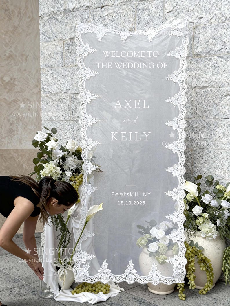 Pode incluir: Uma placa de boas-vindas de casamento com borda de renda branca e o texto "WELCOME TO THE WEDDING OF AXEL and KEILY". A data "18.10.2025" e o local "Peekskill, NY" tamb&eacute;m s&atilde;o exibidos. A placa &eacute; decorada com flores brancas e vegeta&ccedil;&atilde;o.