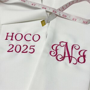 May include: White fabric with embroidered magenta text reading "HOCO 2025" and a monogram "SNJ". A measuring tape is draped across the fabric, showing measurements up to 52 inches.