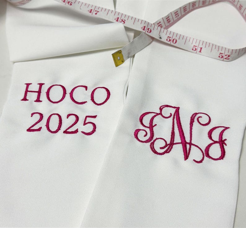 May include: White fabric with embroidered magenta text reading "HOCO 2025" and a monogram "SNJ". A measuring tape is draped across the fabric, showing measurements up to 52 inches.
