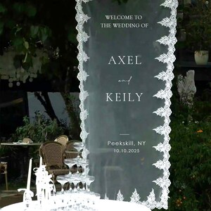 Pode incluir: Uma placa de boas-vindas de casamento branca transparente com borda de renda decorativa. A placa diz "Bem-vindos ao casamento de Axel e Keily" e inclui a data e o local, Peekskill, NY 10.10.2025. Uma torre de champanhe est&aacute; em primeiro plano.