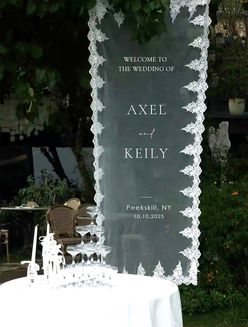 Pode incluir: Uma placa de boas-vindas de casamento branca transparente com borda de renda decorativa. A placa diz "Bem-vindos ao casamento de Axel e Keily" e inclui a data e o local, Peekskill, NY 10.10.2025. Uma torre de champanhe est&aacute; em primeiro plano.