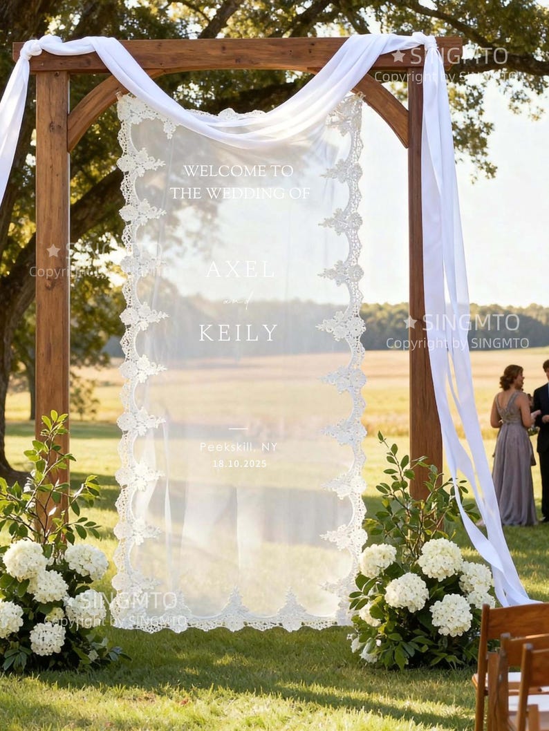 Pode incluir: Um arco de casamento de madeira com um painel de tecido branco transparente com o texto "WELCOME TO THE WEDDING OF AXEL and KEILY" e a data 18.10.2025. O painel &eacute; adornado com uma delicada borda de renda. Tecido branco drapeja do arco, e flores brancas e vegeta&ccedil;&atilde;o est&atilde;o na base.