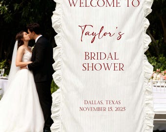 Könnte beinhalten: Weißes Banner mit Rüschenkante und vertikalen Streifen, das in bordeauxfarbenem Text "Welcome to Taylor's Bridal Shower" ankündigt. Das Banner enthält auch Ort und Datum: Dallas, Texas, 15. November 2025. Ein Paar küsst sich im Hintergrund.