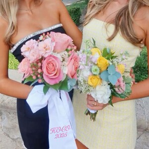 May include: Two women holding bouquets of flowers. One bouquet features pink roses and is paired with a black dress with a jeweled neckline and a white sash with pink text. The other bouquet has yellow and white flowers and is paired with a yellow dress.