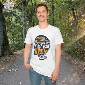 May include: White t-shirt with the text "Always Keep an Open Mind" in a black, orange, and white design. The graphic is shaped like a head. The person is wearing blue jeans and standing on a path in a forest.