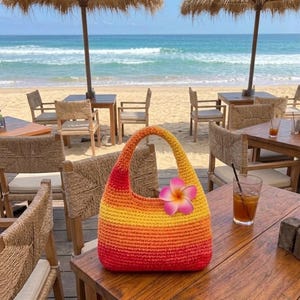 Puede incluir: Un bolso de ganchillo con un patrón de degradado vibrante que transiciona de amarillo en la parte superior a naranja y rojo en la parte inferior. El bolso tiene una sola asa redondeada y está decorado con una flor de plumeria rosa y amarilla.