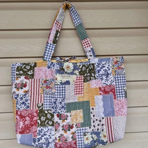 May include: A patchwork tote bag with a variety of fabric patterns, including floral, gingham, and striped designs. The bag has a rectangular shape with a matching patchwork handle. A small label with text is visible on the front.