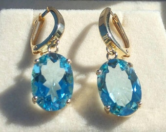 May include: A pair of gold earrings with oval-shaped blue topaz gemstones. The earrings have a simple design and are set in a gold setting.