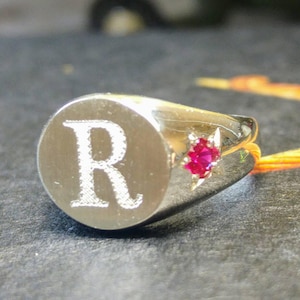May include: A silver signet ring with a raised letter "R" and a small red gemstone.