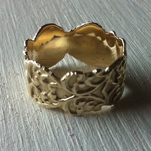 May include: A gold ring with a detailed, intricate design. The ring is wide and has a smooth, polished finish.
