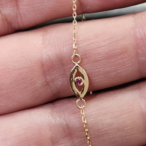 May include: A gold chain bracelet with a small, openwork eye charm featuring a red gemstone in the center.