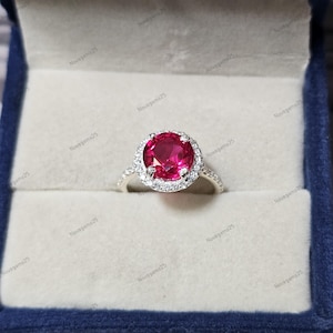 以下が含まれることがあります： 大きな丸いルビーレッドの宝石を、小さな透明な宝石のハローで囲んだシルバーリング。 リングは青いベルベットの箱に入れられ、ジュエリーの輝きを際立たせています。 バンドにも小さな宝石が飾られています。