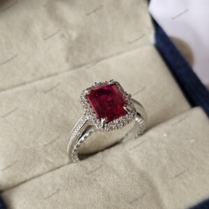 Puede incluir: Un anillo plateado con una gran gema rectangular de color rojo intenso en el centro, rodeada de piedras más pequeñas y transparentes. La banda del anillo tiene detalles intrincados y está en una caja de joyería azul.