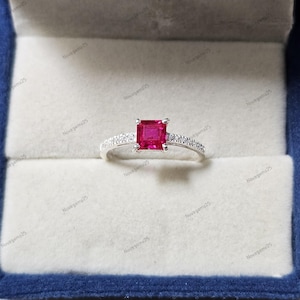 Puede incluir: Un anillo de plata con una gema roja cuadrada y vibrante en el centro, flanqueada por piedras más pequeñas y claras a lo largo de la banda. El anillo se muestra en una caja de joyería azul con interior blanco.
