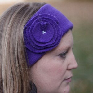 Könnte beinhalten: Ein lilafarbenes Fleece-Stirnband mit einem großen lilafarbenen Blumenakzent. Das Stirnband wird von einer Person mit langen blonden Haaren getragen.
