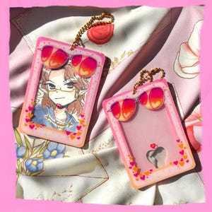 May include: Two pink keychains with a sunglasses design. One keychain features an illustration of a person wearing glasses, while the other has a heart design. Both keychains have a gold chain and a heart-shaped charm.