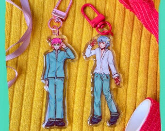 Puede incluir: Dos llaveros de estilo anime con personajes masculinos con trajes turquesa y blancos. Los llaveros tienen cierres plateados. Un personaje tiene el pelo rosa y el otro azul. El fondo es amarillo.