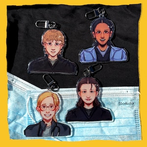 May include: Four acrylic keychains with illustrated portraits of people. Each keychain has a silver clasp. The portraits show individuals with different hairstyles and clothing, set against a black background and a blue medical mask.