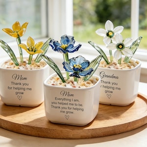 Puede incluir: Tres macetas cuadradas de cerámica blanca con flores de cristal amarillas, azules y blancas. Cada maceta tiene un mensaje para Mamá y Abuela, con un símbolo de corazón. Las macetas están sobre una bandeja de madera.