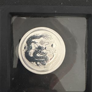 Może przedstawiać: Srebrna moneta z szczeg&oacute;łowym wzorem smoka. Moneta jest oznaczona chińskimi znakami i napisem "Year of the Dragon". Moneta jest umieszczona w przezroczystym plastikowym uchwycie na czarnym tle.