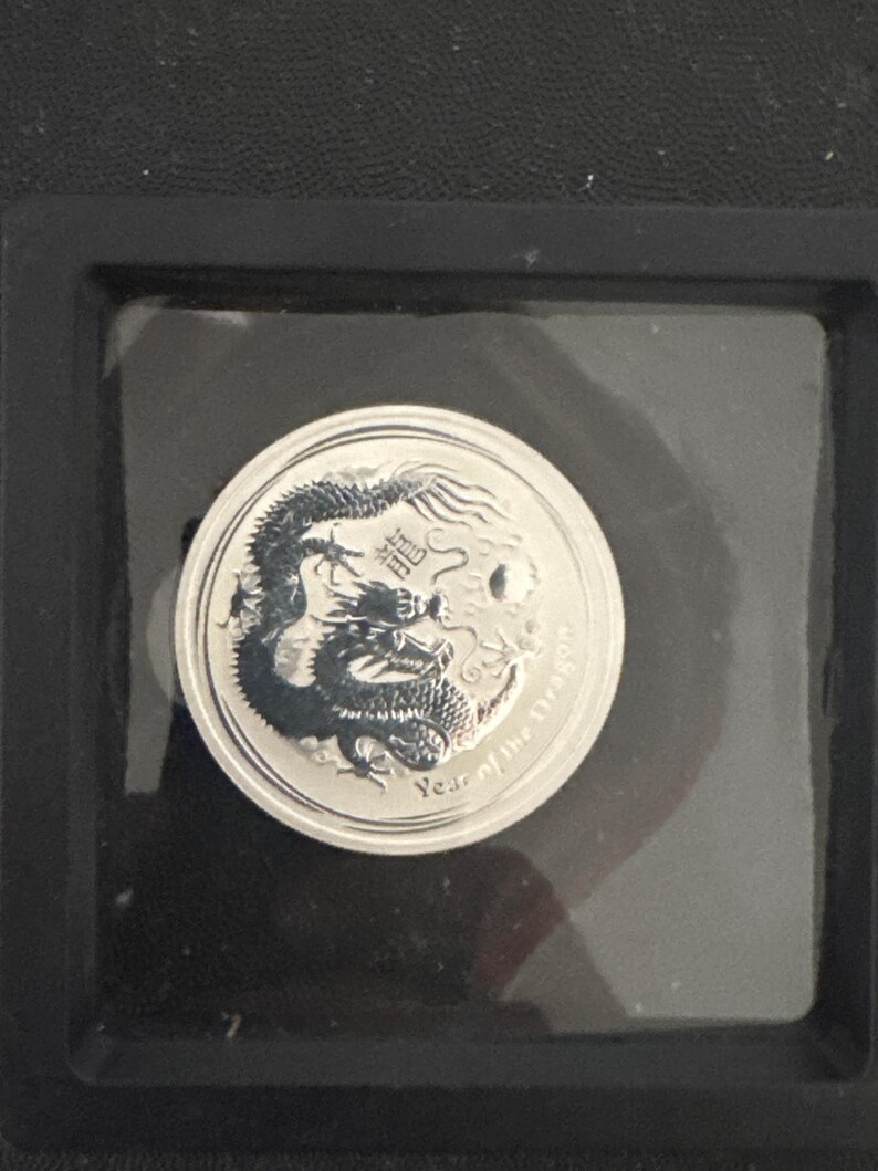 Może przedstawiać: Srebrna moneta z szczeg&oacute;łowym wzorem smoka. Moneta jest oznaczona chińskimi znakami i napisem "Year of the Dragon". Moneta jest umieszczona w przezroczystym plastikowym uchwycie na czarnym tle.