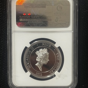 Peut inclure: Une pièce de monnaie en argent de Niue, dans un étui de protection transparent. La pièce représente un profil de la reine Elizabeth II et le texte "TWO DOLLAR" et l'année "2012". L'étui porte une étiquette de Numismatic Guaranty Corporation.