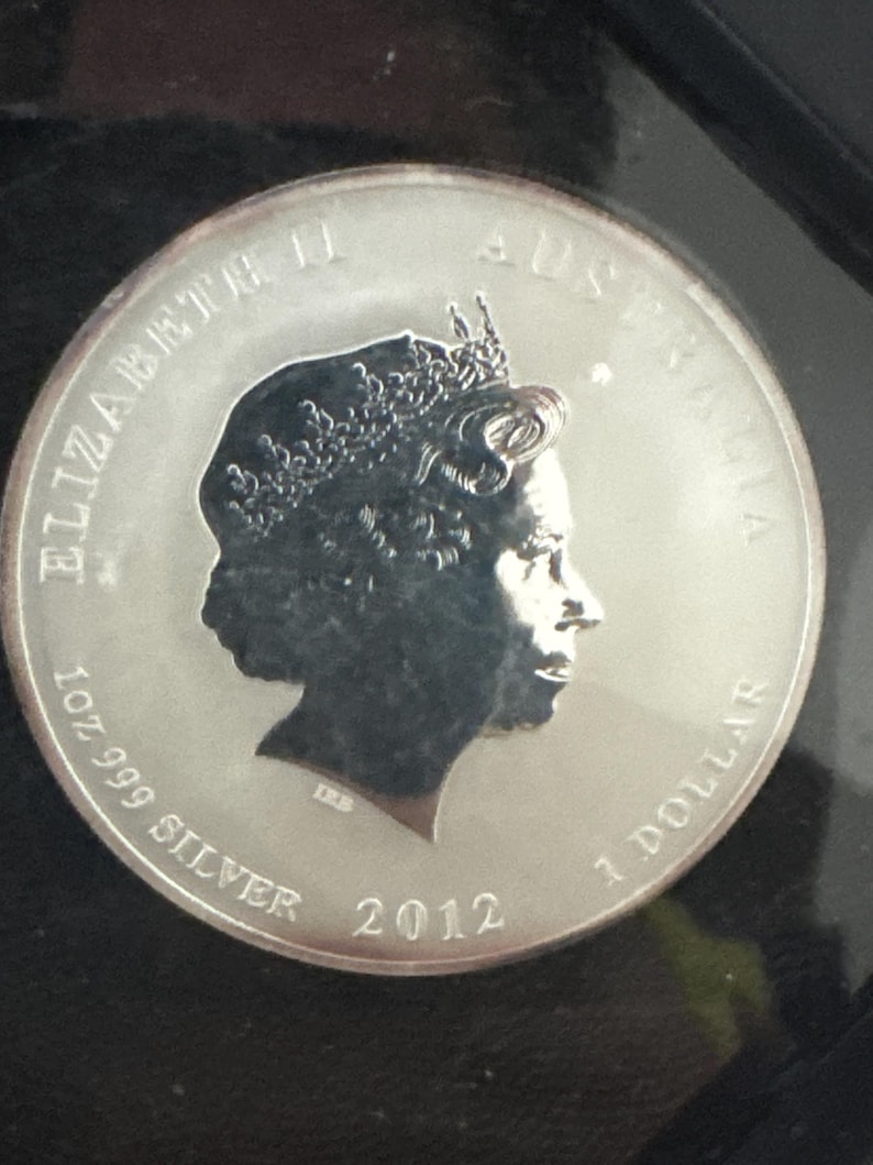 Może przedstawiać: Srebrna australijska moneta 1-dolarowa z 2012 roku. Moneta przedstawia profil kr&oacute;lowej Elżbiety II, napis "ELIZABETH II AUSTRALIA", "1 OZ 999 SILVER" i "1 DOLLAR".