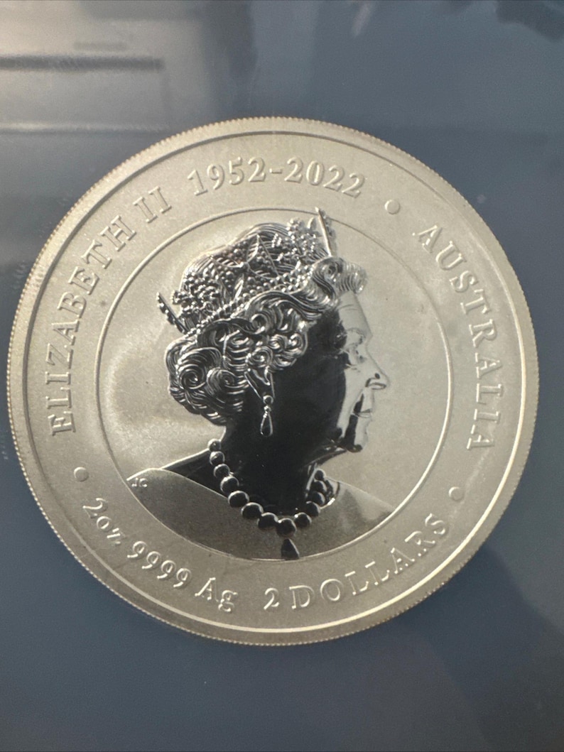 Może przedstawiać: Srebrna moneta z profilem ukoronowanej postaci, z napisem "ELIZABETH II 1952-2022 AUSTRALIA". Moneta zawiera r&oacute;wnież napis "2oz 9999 Ag 2 DOLLARS".