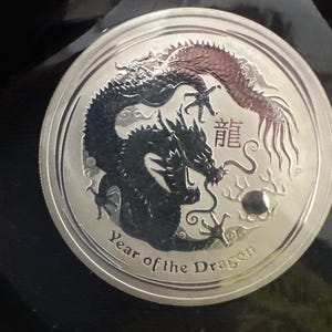 Może przedstawiać: Srebrna moneta z szczeg&oacute;łowym przedstawieniem dw&oacute;ch smok&oacute;w, jeden czarny i jeden czerwony. Moneta jest podpisana "Year of the Dragon" i chińskimi znakami. Moneta ma błyszczącą, odblaskową powierzchnię.