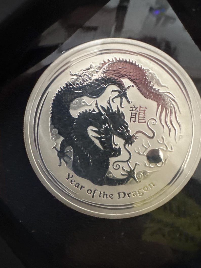 Może przedstawiać: Srebrna moneta z szczeg&oacute;łowym przedstawieniem dw&oacute;ch smok&oacute;w, jeden czarny i jeden czerwony. Moneta jest podpisana "Year of the Dragon" i chińskimi znakami. Moneta ma błyszczącą, odblaskową powierzchnię.