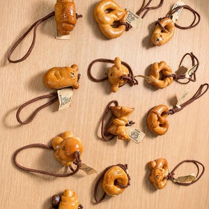 May include: A collection of eleven hair ties featuring miniature, glossy, golden-brown bread-shaped charms in various forms including croissants, pretzels, and animal shapes. Each hair tie has a brown elastic band and a small beige paper tag.