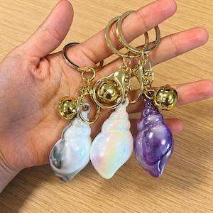 May include: Three seashell-shaped keychains featuring gold-tone metal rings and lobster clasps with attached gold bells. The shells have a smooth, glossy finish in white with grey marbling, iridescent pearlescent white, and deep purple marble.