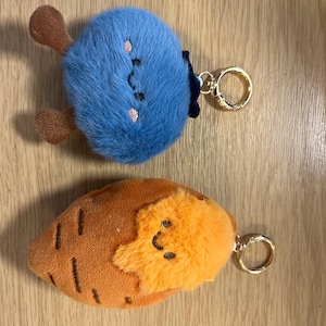 May include: Two plush keychains featuring embroidered faces. The top keychain is a round, fuzzy blue character with small brown limbs and a gold-toned metal ring. The bottom keychain is a brown, carrot-shaped character with a fuzzy orange top.