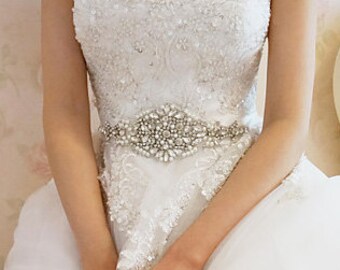 Op de afbeelding: Een witte off-the-shoulder trouwjurk met ingewikkeld kant en kralenwerk. De jurk heeft een sprankelende, versierde riem en een volle rok. De bruid zit en houdt een roze bloem vast.